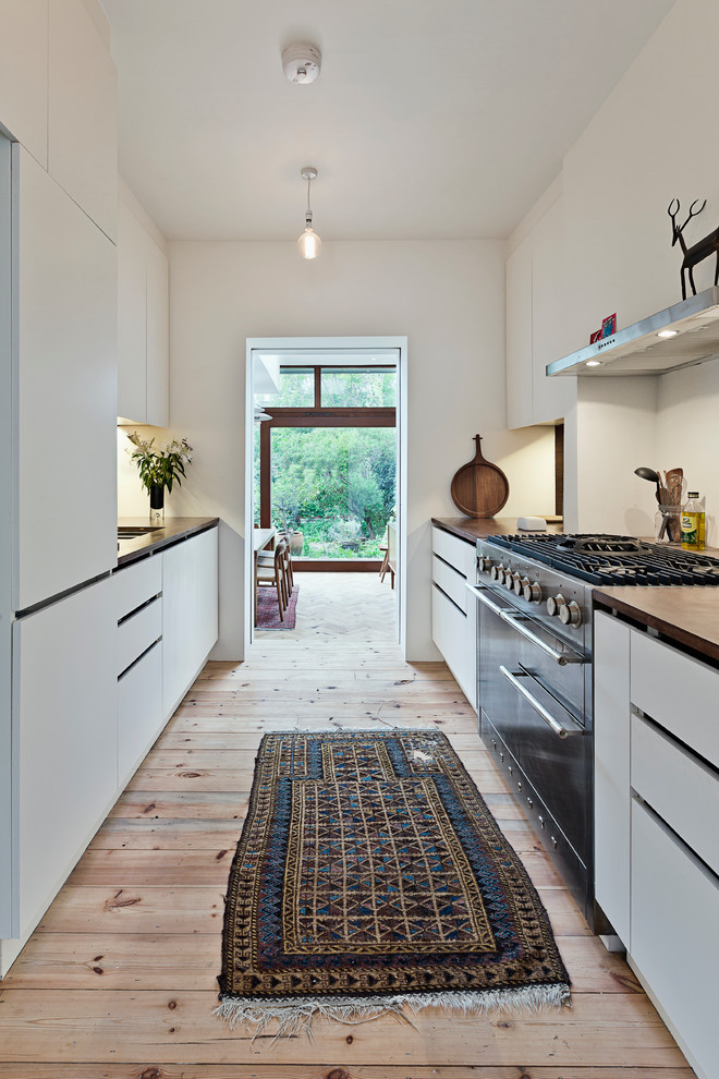 Mid-sized contemporary galley separate kitchen in Cambridgeshire with flat-panel cabinets, white cabinets, stainless steel appliances, light hardwood floors, a drop-in sink, wood benchtops and white splashback.