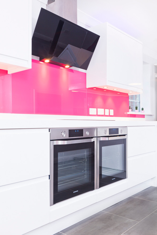 This is an example of a mid-sized contemporary galley eat-in kitchen in Buckinghamshire with flat-panel cabinets, white cabinets, solid surface benchtops, pink splashback, glass sheet splashback, stainless steel appliances, vinyl floors, a peninsula and an integrated sink.