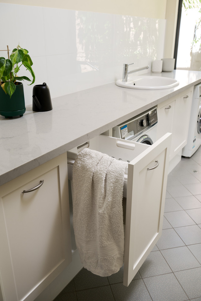 This is an example of a mid-sized traditional single-wall dedicated laundry room in Perth with an utility sink, shaker cabinets, white cabinets, quartz benchtops, white splashback, porcelain splashback, white walls, porcelain floors, an integrated washer and dryer, grey floor and white benchtop.