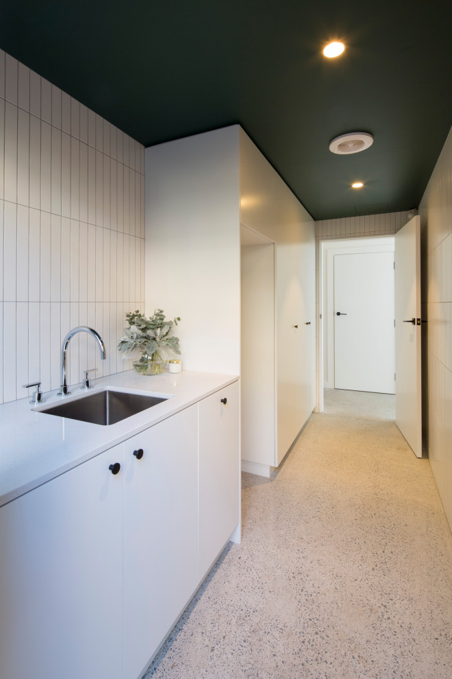 Mid-sized transitional single-wall utility room in Sydney with an undermount sink, white cabinets, white walls, concrete floors, a stacked washer and dryer, grey floor and white benchtop.