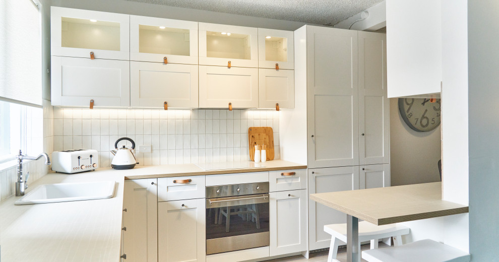Inspiration for a small beach style l-shaped kitchen pantry in Melbourne with a farmhouse sink, shaker cabinets, white cabinets, laminate benchtops, white splashback, ceramic splashback, stainless steel appliances, vinyl floors, a peninsula, grey floor and beige benchtop.