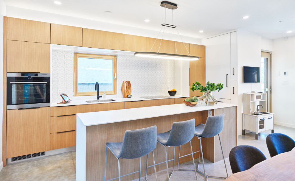 Photo of a mid-sized contemporary l-shaped eat-in kitchen in Vancouver with an undermount sink, flat-panel cabinets, light wood cabinets, quartz benchtops, white splashback, mosaic tile splashback, panelled appliances, concrete floors, with island, grey floor and white benchtop.