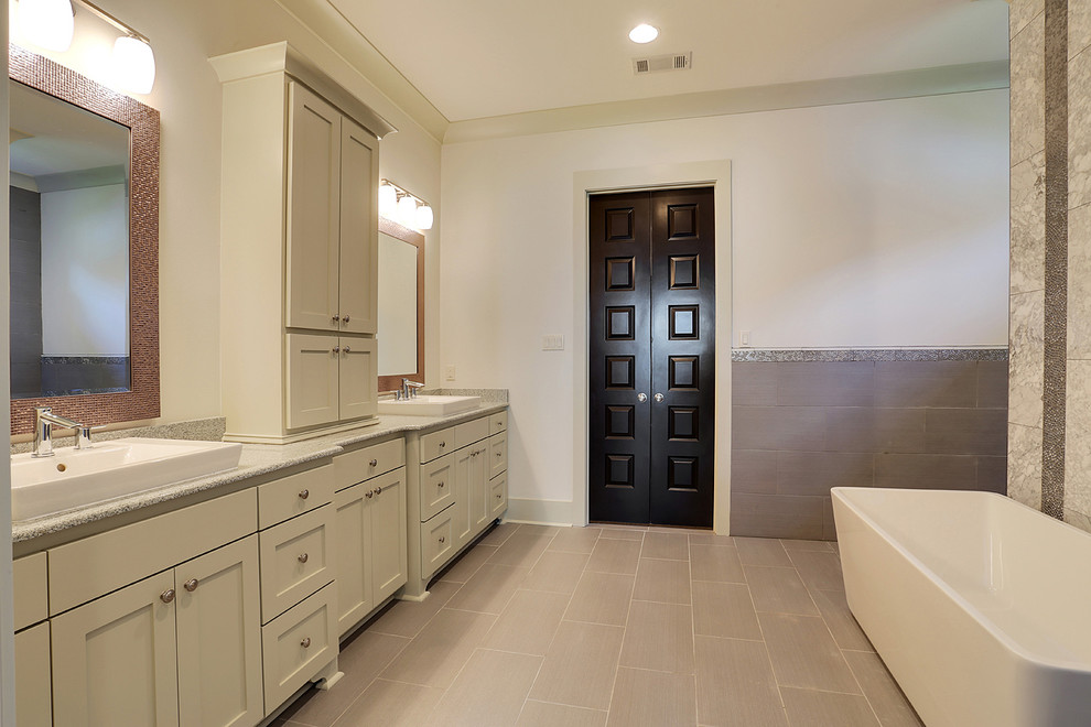 Photo of a large transitional master bathroom in New Orleans with raised-panel cabinets, white cabinets, a freestanding tub, an open shower, a one-piece toilet, gray tile, multi-coloured tile, white tile, ceramic tile, white walls, ceramic floors, a drop-in sink and glass benchtops.
