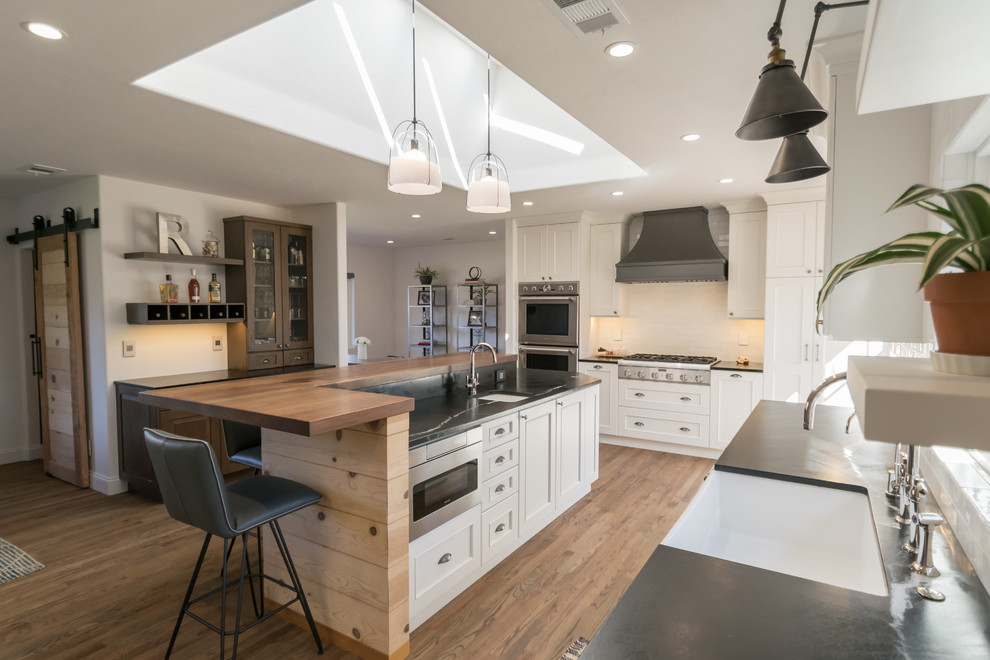 Open concept kitchen - huge transitional l-shaped medium tone wood floor and brown floor open concept kitchen idea in San Francisco with a farmhouse sink, recessed-panel cabinets, white cabinets, soapstone countertops, white backsplash, stainless steel appliances, an island and black countertops