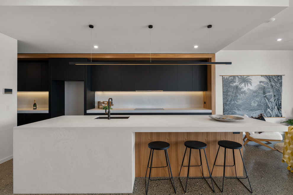 This is an example of a beach style galley open plan kitchen in Sunshine Coast with an undermount sink, flat-panel cabinets, black cabinets, white splashback, black appliances, terrazzo floors, with island, brown floor and white benchtop.