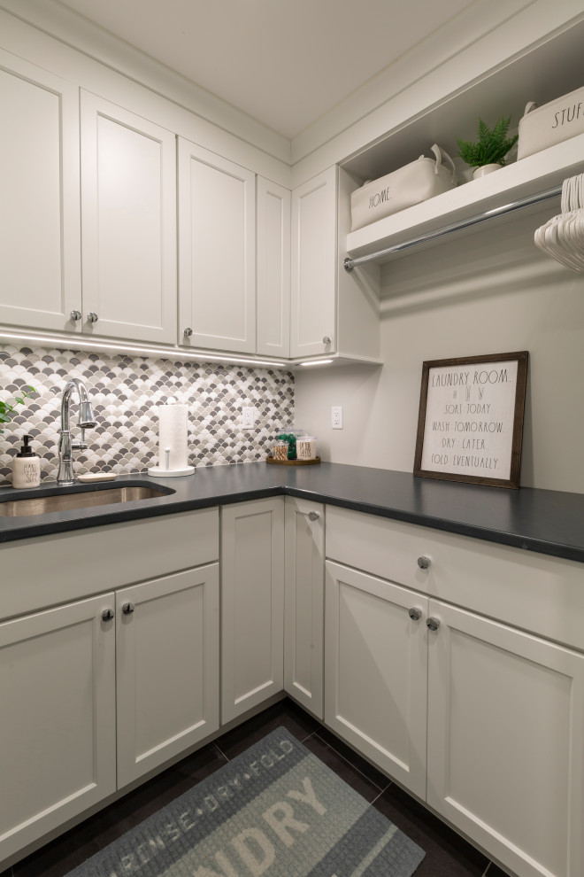 Inspiration for a mid-sized transitional l-shaped dedicated laundry room in New York with an undermount sink, shaker cabinets, white cabinets, quartz benchtops, multi-coloured splashback, mosaic tile splashback, beige walls, a stacked washer and dryer and black benchtop.