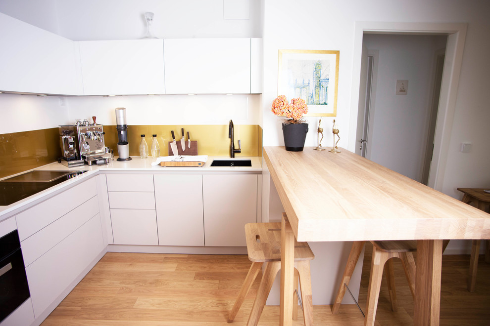 This is an example of a small modern l-shaped eat-in kitchen in Munich with an undermount sink, flat-panel cabinets, beige cabinets, glass sheet splashback, black appliances, medium hardwood floors, a peninsula, brown floor and white benchtop.