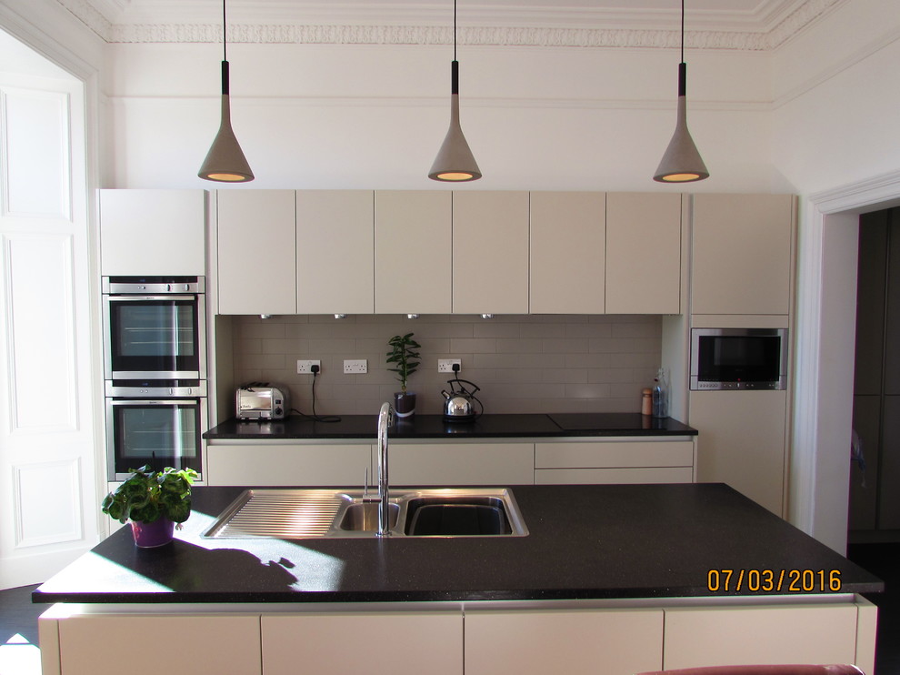 This is an example of a mid-sized modern single-wall eat-in kitchen in Edinburgh with an undermount sink, flat-panel cabinets, beige cabinets, solid surface benchtops, grey splashback, porcelain splashback, stainless steel appliances, vinyl floors and with island.
