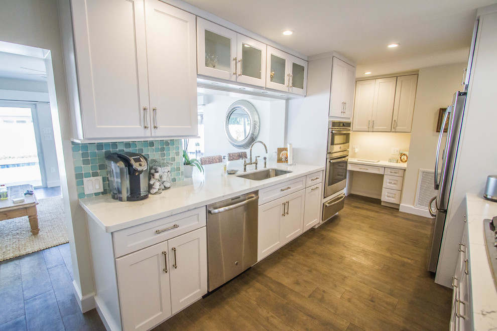 Large beach style galley kitchen in Miami with an undermount sink, shaker cabinets, white cabinets, quartzite benchtops, blue splashback, glass tile splashback, stainless steel appliances, dark hardwood floors, a peninsula, brown floor and white benchtop.