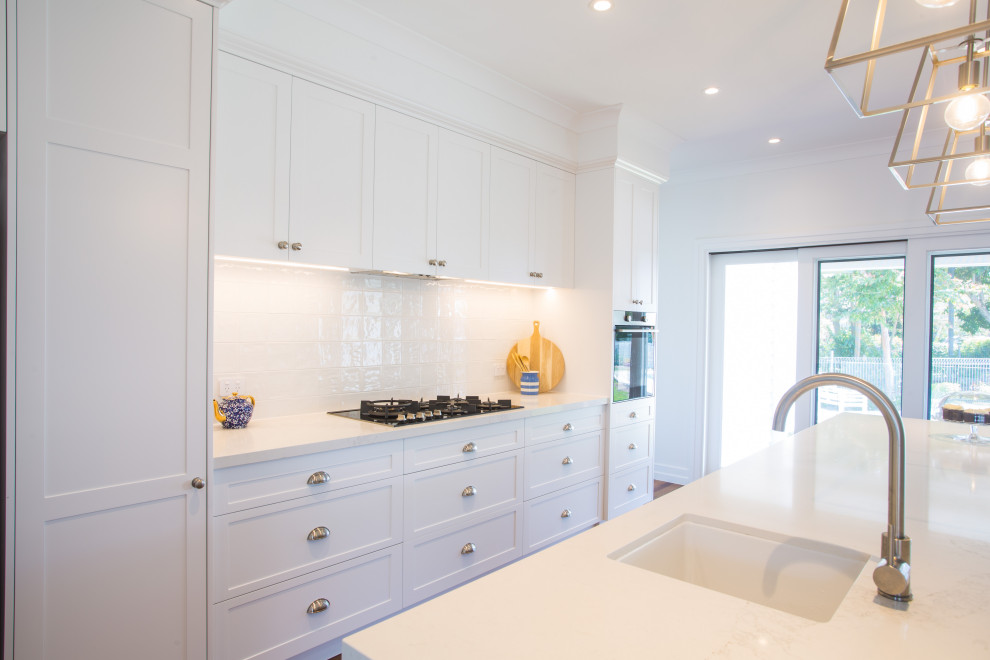This is an example of a large galley kitchen pantry in Brisbane with a single-bowl sink, shaker cabinets, blue cabinets, engineered stone countertops, white splashback, ceramic splashback, black appliances, dark hardwood flooring, an island, brown floors and white worktops.
