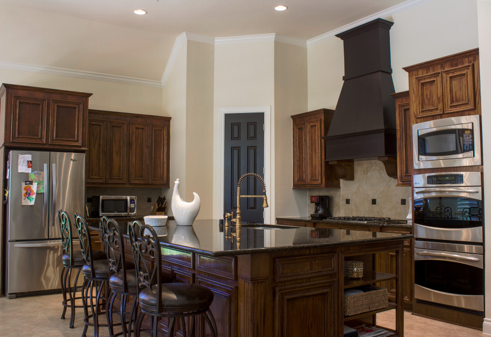 This is an example of a large country u-shaped eat-in kitchen in Houston with an undermount sink, raised-panel cabinets, medium wood cabinets, granite benchtops, beige splashback, travertine splashback, stainless steel appliances, ceramic floors, with island, beige floor, black benchtop and coffered.