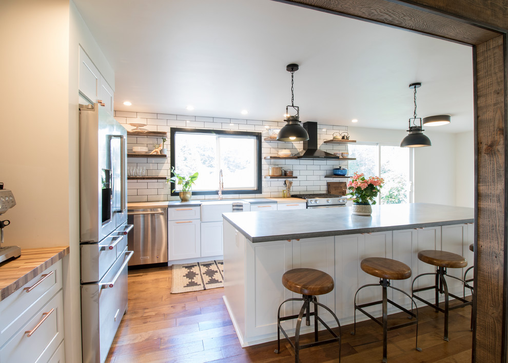 Photo of a mid-sized transitional l-shaped eat-in kitchen in Seattle with a farmhouse sink, shaker cabinets, white cabinets, concrete benchtops, white splashback, ceramic splashback, stainless steel appliances, medium hardwood floors, with island, brown floor and grey benchtop.