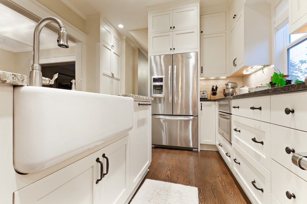 This is an example of a traditional u-shaped open plan kitchen in Seattle with a farmhouse sink, shaker cabinets, white cabinets, granite benchtops, white splashback, ceramic splashback and stainless steel appliances.