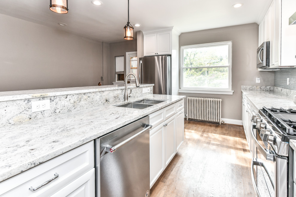 Large modern l-shaped eat-in kitchen in DC Metro with a double-bowl sink, raised-panel cabinets, white cabinets, granite benchtops, blue splashback, glass tile splashback, stainless steel appliances, medium hardwood floors, with island, brown floor and grey benchtop.