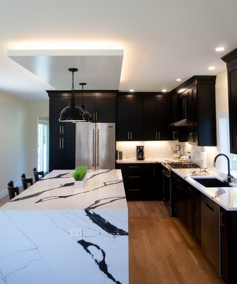 This is an example of a large modern l-shaped eat-in kitchen in Boston with shaker cabinets, black cabinets, quartz benchtops, white splashback, glass tile splashback, stainless steel appliances, light hardwood floors, with island, brown floor and white benchtop.