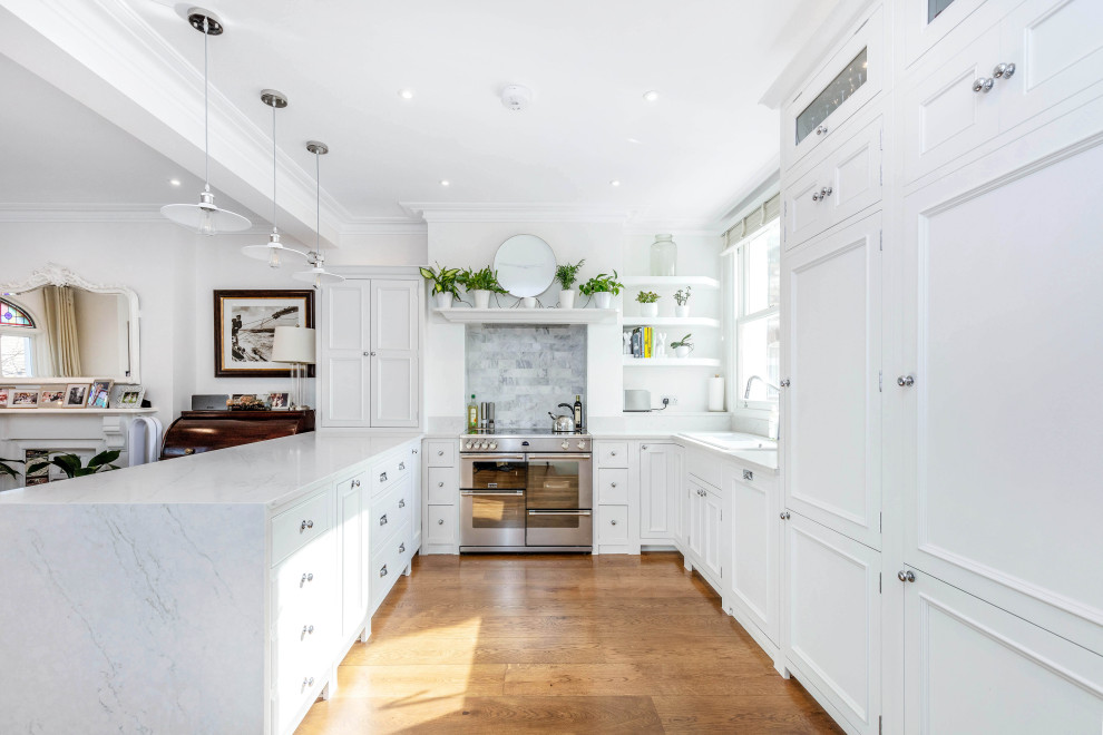 Mid-sized transitional u-shaped open plan kitchen in London with a drop-in sink, shaker cabinets, white cabinets, marble benchtops, white splashback, marble splashback, panelled appliances, light hardwood floors, with island, brown floor and white benchtop.