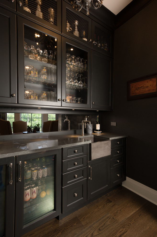 This is an example of a mid-sized traditional single-wall wet bar in Chicago with an integrated sink, glass-front cabinets, dark wood cabinets, quartz benchtops, mirror splashback, dark hardwood floors, brown floor and grey benchtop.