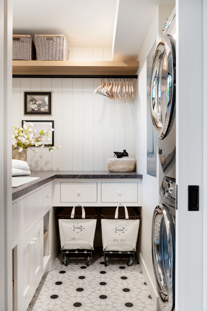 Inspiration for a beach style l-shaped dedicated laundry room in Minneapolis with a drop-in sink, white cabinets, white walls, a stacked washer and dryer, multi-coloured floor, black benchtop and planked wall panelling.
