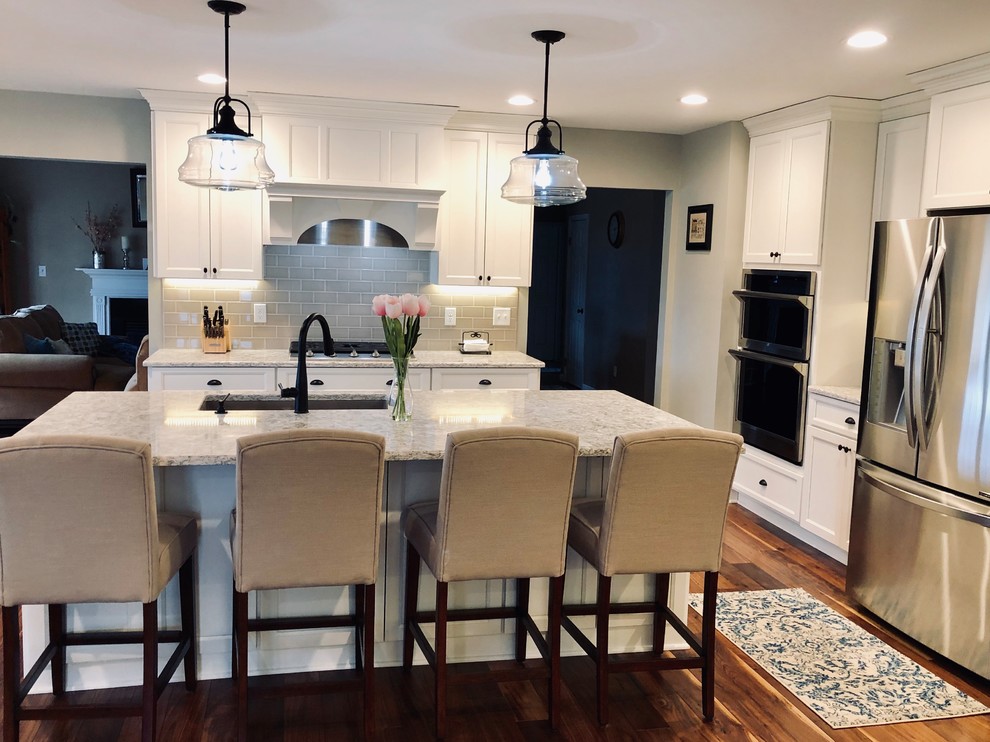 This is an example of an expansive country eat-in kitchen in New York with an undermount sink, shaker cabinets, white cabinets, quartz benchtops, white splashback, ceramic splashback, stainless steel appliances, medium hardwood floors, with island, multi-coloured floor and white benchtop.