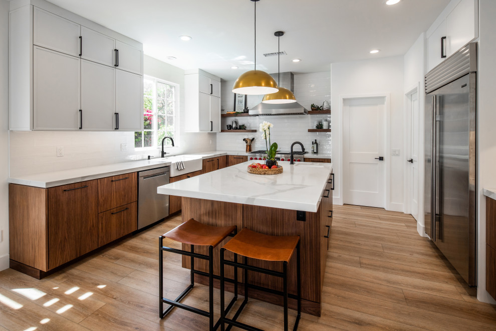 Example of a mid-sized 1950s u-shaped laminate floor and beige floor open concept kitchen design in San Diego with a farmhouse sink, flat-panel cabinets, medium tone wood cabinets, marble countertops, white backsplash, ceramic backsplash, stainless steel appliances, an island and white countertops