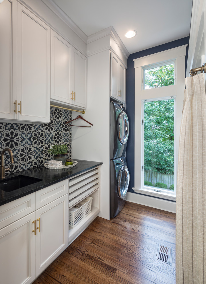 Photo of a transitional single-wall dedicated laundry room in Cincinnati with an undermount sink, recessed-panel cabinets, white cabinets, blue walls, dark hardwood floors, a stacked washer and dryer, brown floor and black benchtop.