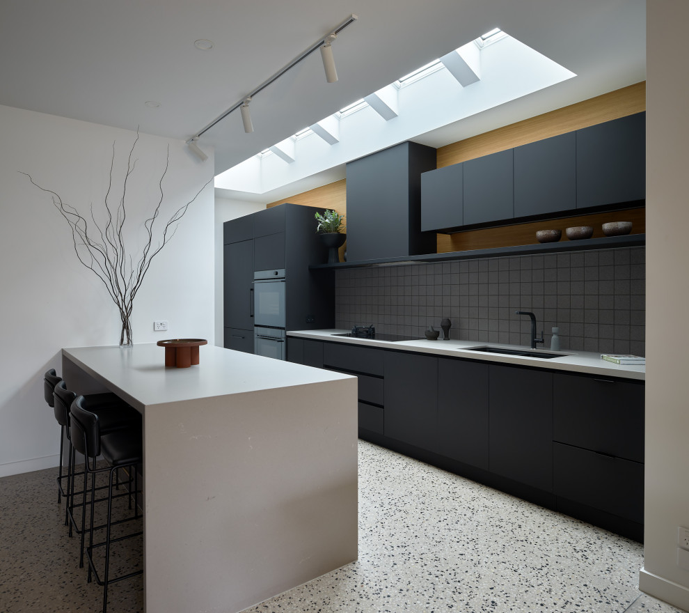 This is an example of a large contemporary kitchen in Melbourne with flat-panel cabinets, black cabinets, solid surface benchtops, grey splashback, mosaic tile splashback, black appliances, concrete floors, with island, multi-coloured floor and grey benchtop.