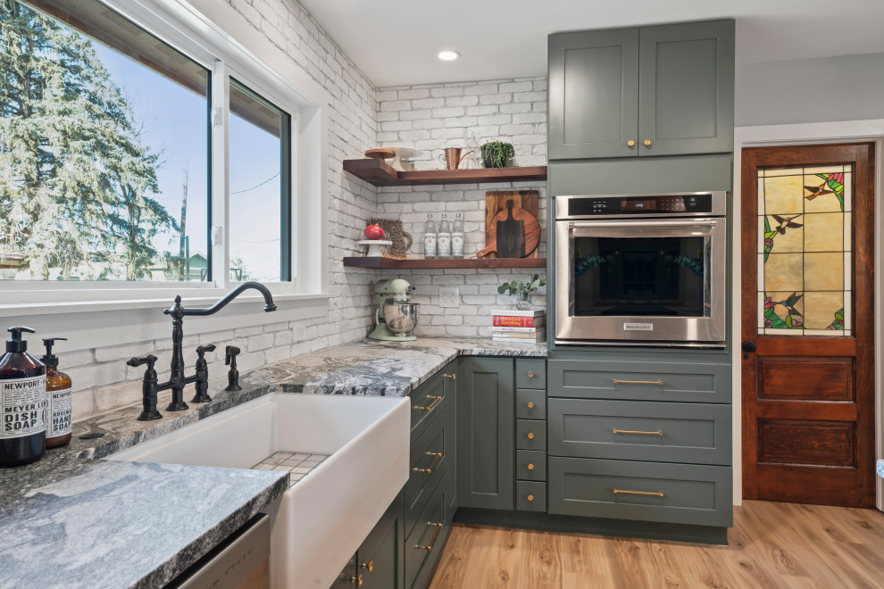 Photo of a large country l-shaped eat-in kitchen in Denver with a farmhouse sink, shaker cabinets, green cabinets, granite benchtops, white splashback, brick splashback, stainless steel appliances, vinyl floors, with island, beige floor, grey benchtop and recessed.