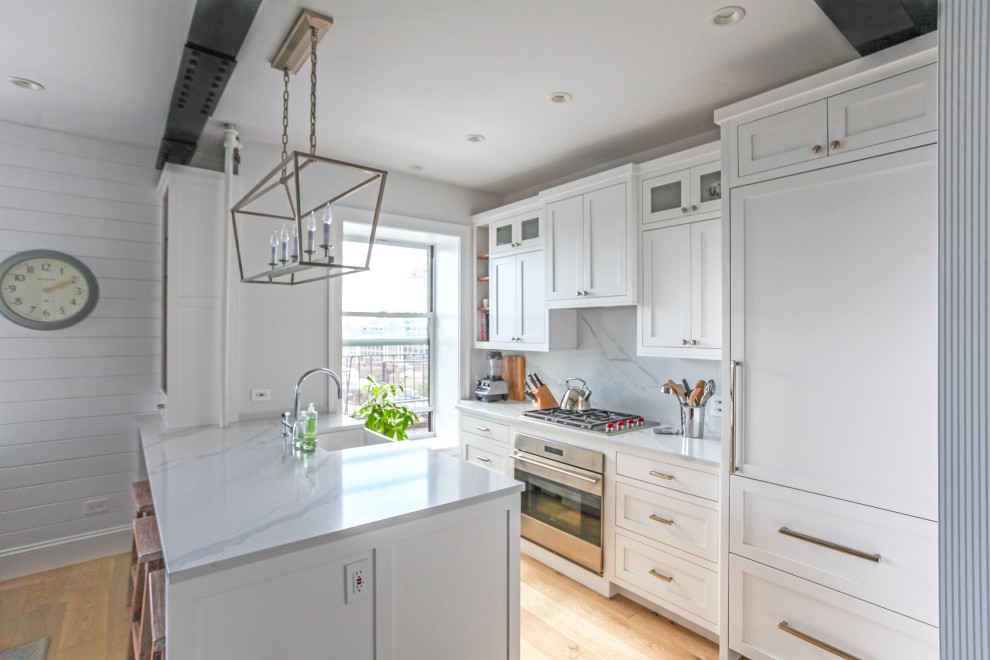 This is an example of a small beach style single-wall eat-in kitchen in New York with a farmhouse sink, recessed-panel cabinets, white cabinets, quartz benchtops, white splashback, engineered quartz splashback, stainless steel appliances, light hardwood floors, a peninsula and white benchtop.