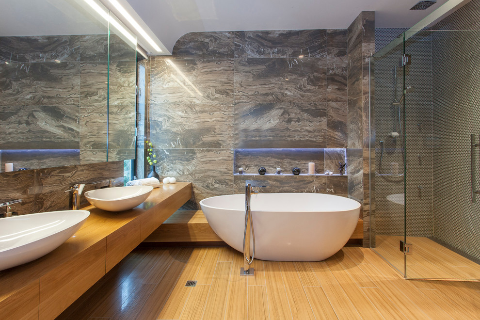 Photo of a contemporary master bathroom in Christchurch with flat-panel cabinets, medium wood cabinets, a freestanding tub, a curbless shower, gray tile, mosaic tile, brown walls, a vessel sink, wood benchtops, medium hardwood floors, a hinged shower door and brown benchtops.