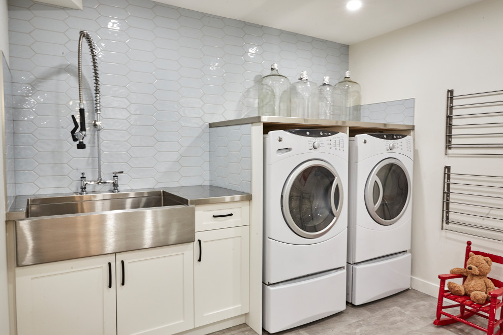 Large modern galley utility room in Edmonton with vinyl floors, grey walls, grey floor, a farmhouse sink, shaker cabinets, white cabinets, stainless steel benchtops, blue splashback, ceramic splashback, a stacked washer and dryer and grey benchtop.