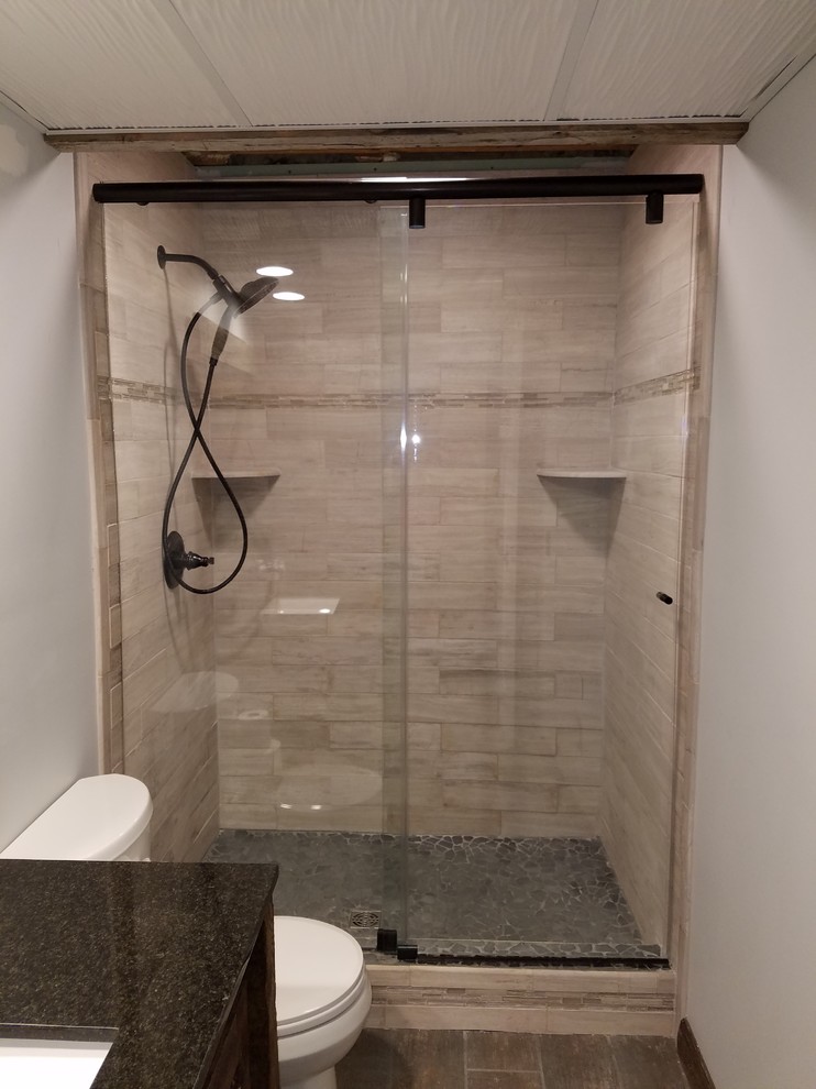 Photo of a mid-sized country 3/4 bathroom in Minneapolis with louvered cabinets, dark wood cabinets, an alcove shower, a two-piece toilet, beige tile, stone tile, grey walls, medium hardwood floors, an undermount sink, engineered quartz benchtops, brown floor and a sliding shower screen.