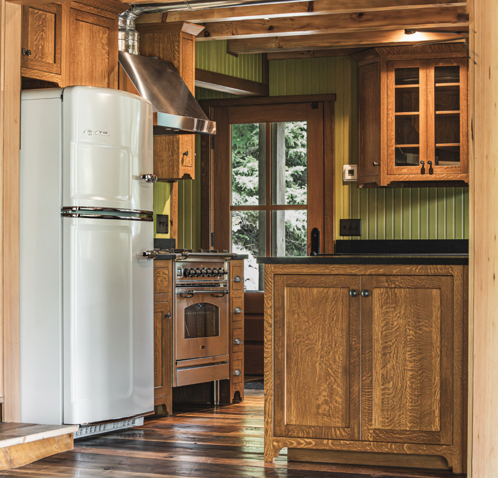 Mountain style medium tone wood floor and exposed beam kitchen photo with an undermount sink, shaker cabinets, granite countertops, black backsplash, granite backsplash, stainless steel appliances, an island and black countertops