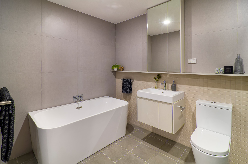 Design ideas for a mid-sized contemporary master bathroom in Sydney with flat-panel cabinets, beige cabinets, a freestanding tub, a one-piece toilet, gray tile, ceramic tile, beige walls, ceramic floors, an integrated sink, laminate benchtops, brown floor and white benchtops.