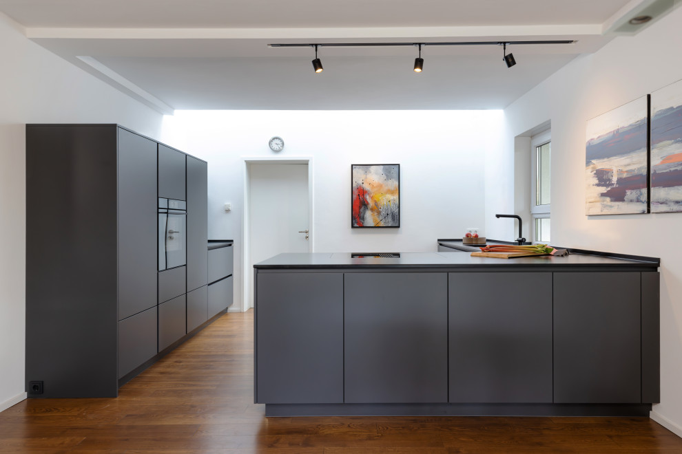 This is an example of a mid-sized contemporary galley open plan kitchen in Frankfurt with an integrated sink, flat-panel cabinets, grey cabinets, granite benchtops, a peninsula and black benchtop.