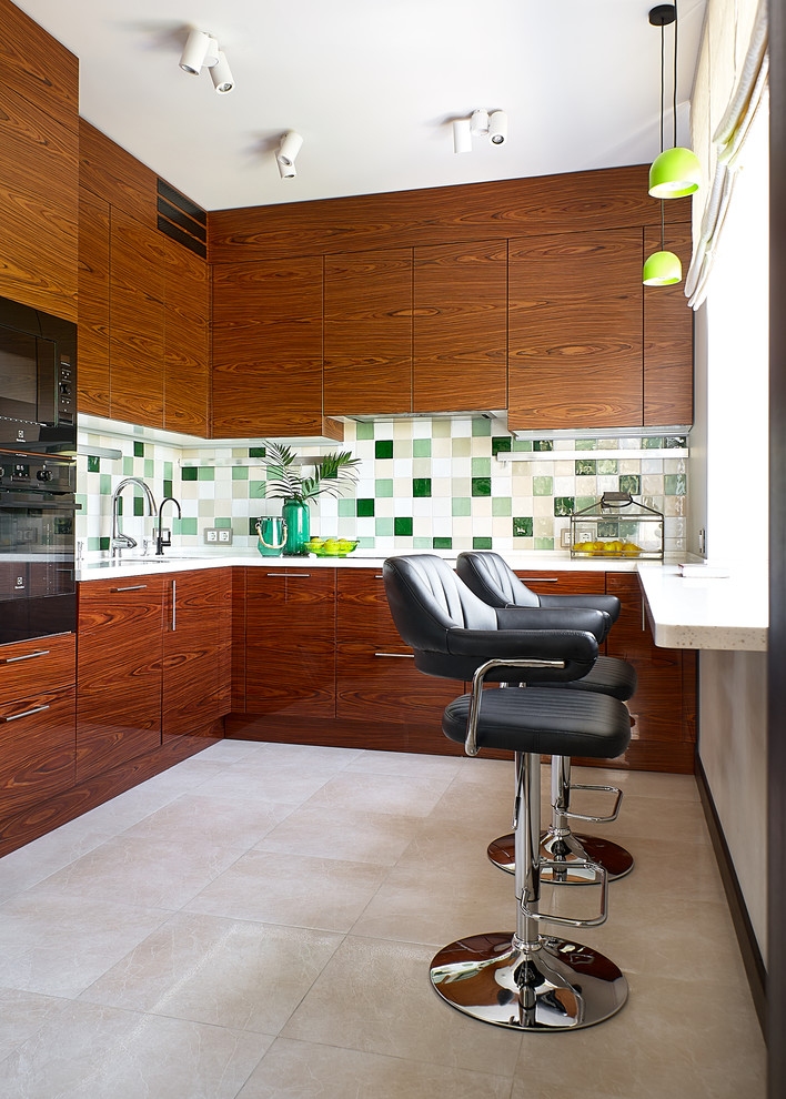 Стильный дизайн: угловая кухня в современном стиле с плоскими фасадами, фасадами цвета дерева среднего тона, зеленым фартуком, черной техникой, бежевым полом и белой столешницей - последний тренд