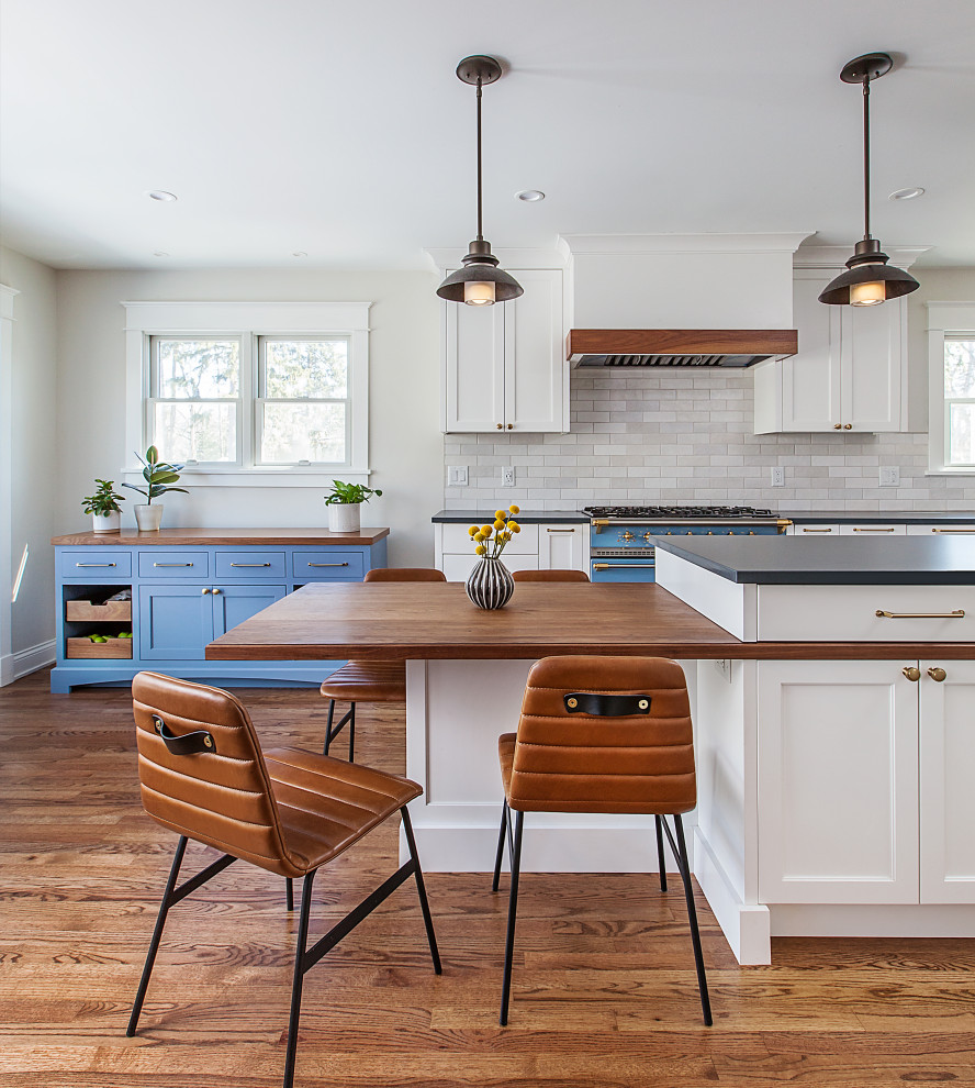 Eat-in kitchen - mid-sized transitional l-shaped medium tone wood floor and brown floor eat-in kitchen idea in Detroit with a farmhouse sink, flat-panel cabinets, white cabinets, quartz countertops, white backsplash, ceramic backsplash, stainless steel appliances, an island and black countertops