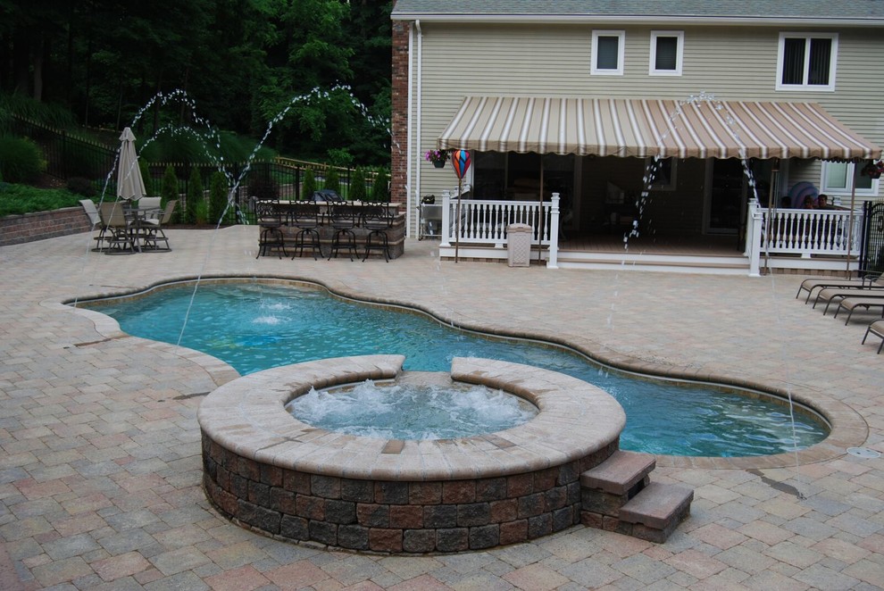 Mid-sized traditional backyard custom-shaped pool in New York with a hot tub and brick pavers.