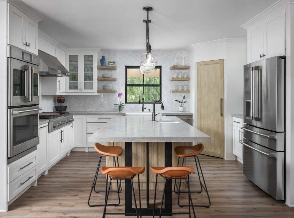 Large eclectic u-shaped kitchen pantry in Orange County with a farmhouse sink, raised-panel cabinets, white cabinets, quartzite benchtops, grey splashback, mosaic tile splashback, stainless steel appliances, medium hardwood floors, with island, brown floor and grey benchtop.