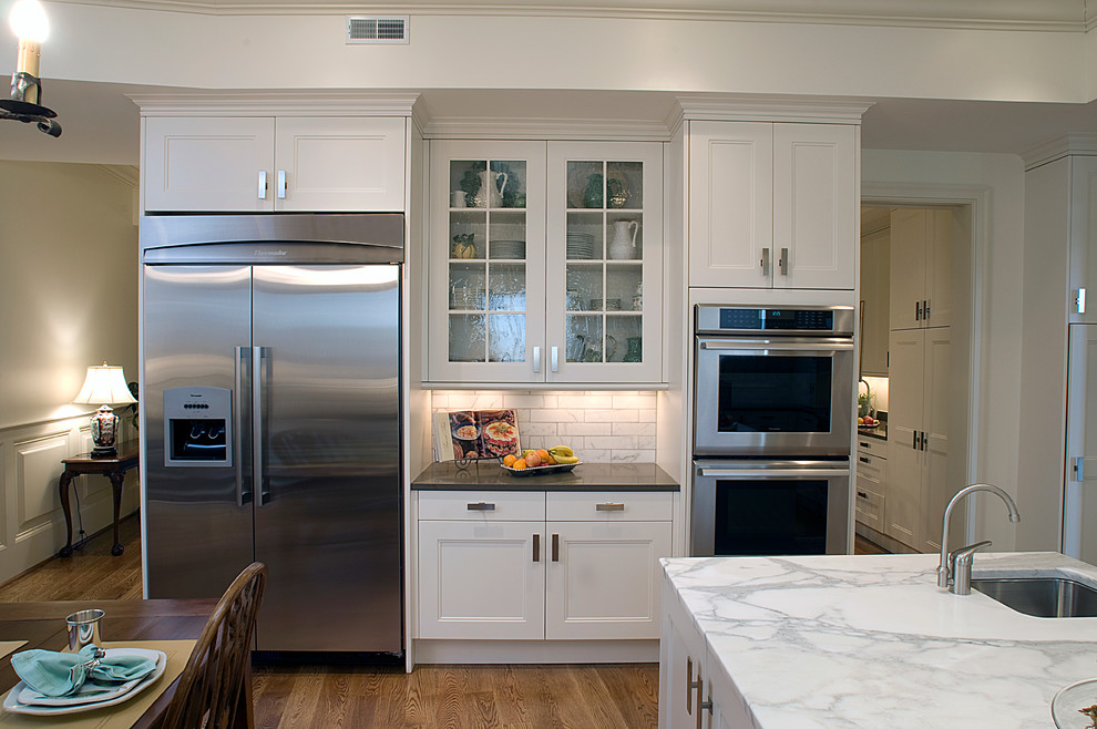 Inspiration for a large transitional u-shaped medium tone wood floor enclosed kitchen remodel with an undermount sink, recessed-panel cabinets, white cabinets, marble countertops, white backsplash, stone tile backsplash, stainless steel appliances and an island