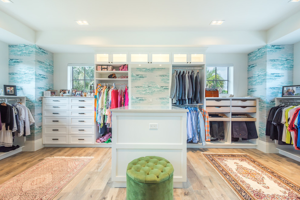This is an example of a tropical gender-neutral walk-in wardrobe in Miami with white cabinets and light hardwood floors.
