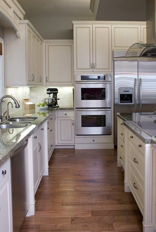 Photo of a traditional kitchen in Los Angeles with stainless steel appliances, a triple-bowl sink, raised-panel cabinets, white cabinets, white splashback and granite benchtops.