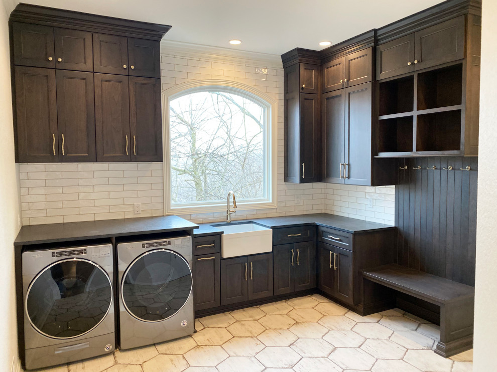 This is an example of a large transitional l-shaped dedicated laundry room in Milwaukee with a farmhouse sink, recessed-panel cabinets, brown cabinets, granite benchtops, white splashback, subway tile splashback, terra-cotta floors, a side-by-side washer and dryer, white floor and black benchtop.