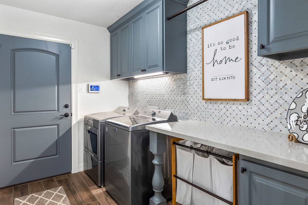 Large transitional single-wall laundry cupboard in Houston with raised-panel cabinets, blue cabinets, grey walls, a side-by-side washer and dryer, brown floor and grey benchtop.