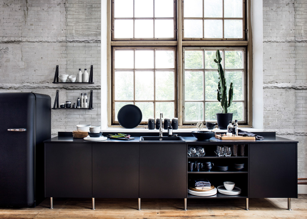 Photo of a mid-sized industrial single-wall open plan kitchen in Gothenburg with a single-bowl sink, flat-panel cabinets, black cabinets, laminate benchtops, black appliances, light hardwood floors, no island, beige floor and black benchtop.