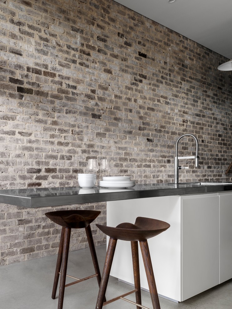 This is an example of a small industrial single-wall eat-in kitchen in Sydney with a single-bowl sink, flat-panel cabinets, white cabinets, stainless steel benchtops, metallic splashback, mirror splashback, stainless steel appliances, concrete floors and with island.