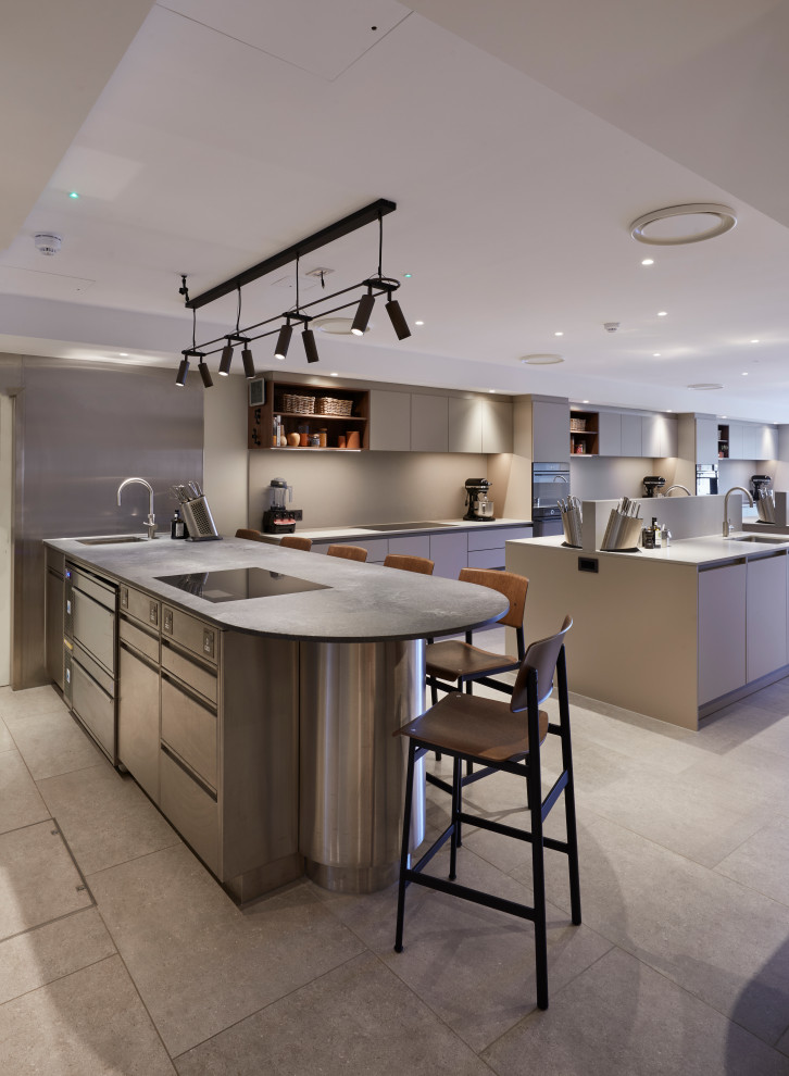 Photo of an expansive contemporary grey and brown galley open plan kitchen in Berkshire with a single-bowl sink, flat-panel cabinets, beige cabinets, concrete worktops, grey splashback, engineered quartz splashback, black appliances, porcelain flooring, multiple islands, beige floors and grey worktops.