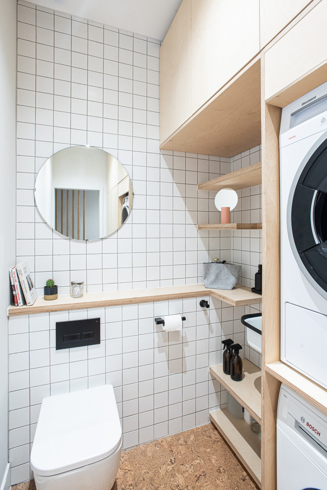 Small scandinavian galley utility room in London with an utility sink, flat-panel cabinets, light wood cabinets, wood benchtops, white splashback, ceramic splashback, white walls, cork floors, a stacked washer and dryer and brown floor.