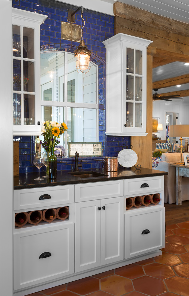 This is an example of a beach style single-wall wet bar in Charlotte with an undermount sink, glass-front cabinets, white cabinets, blue splashback, subway tile splashback, terra-cotta floors, orange floor and black benchtop.