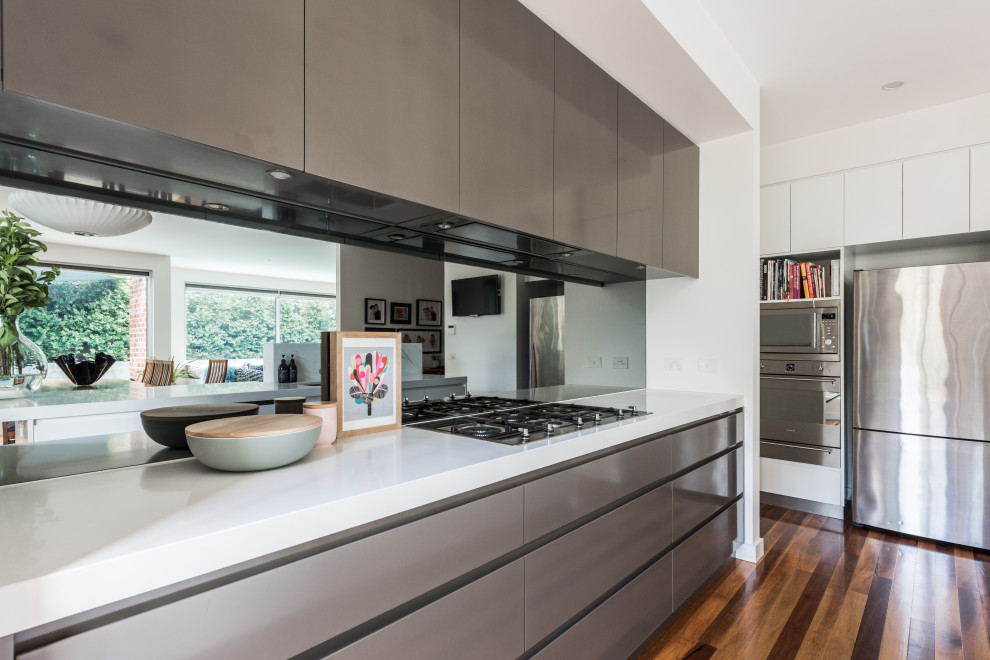 This is an example of a contemporary kitchen in Melbourne with flat-panel cabinets, grey cabinets, metallic splashback, mirror splashback, stainless steel appliances, dark hardwood floors, brown floor and white benchtop.