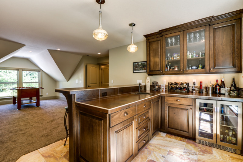 Mid-sized country l-shaped wet bar in Portland with a drop-in sink, raised-panel cabinets, dark wood cabinets, tile benchtops, marble floors, multi-coloured splashback, glass tile splashback and beige floor.